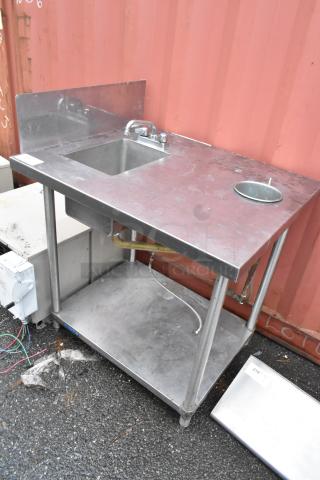 Stainless steel commercial sink with faucet, side drain, and lower shelf. Some wear visible, ideal for kitchen use.