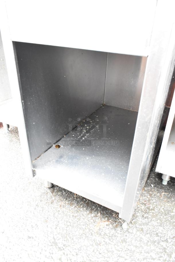 Stainless steel open cabinet with lower shelf, visible wear and markings, resting on adjustable feet.