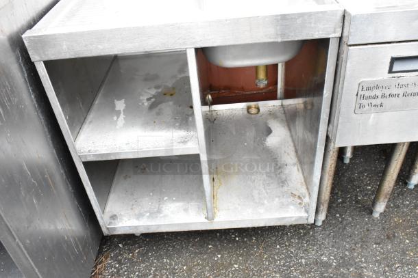 Stainless steel utility sink with open storage, worn condition, visible plumbing, and side label advising handwashing.