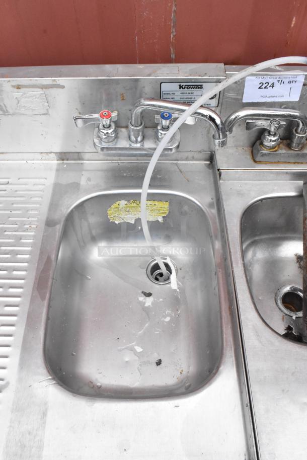 Stainless steel sink with dual faucets and a labeled Krowne model tag. Visible wear, with a taped message and attached tube.