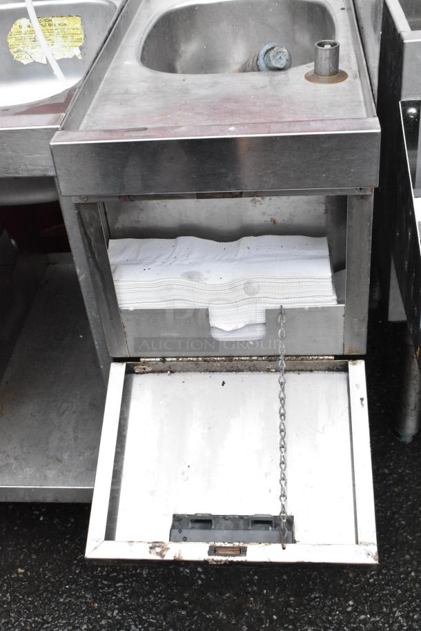 Stainless steel sink unit with built-in paper towel dispenser. Visible wear and rust. Yellow caution label on the side.