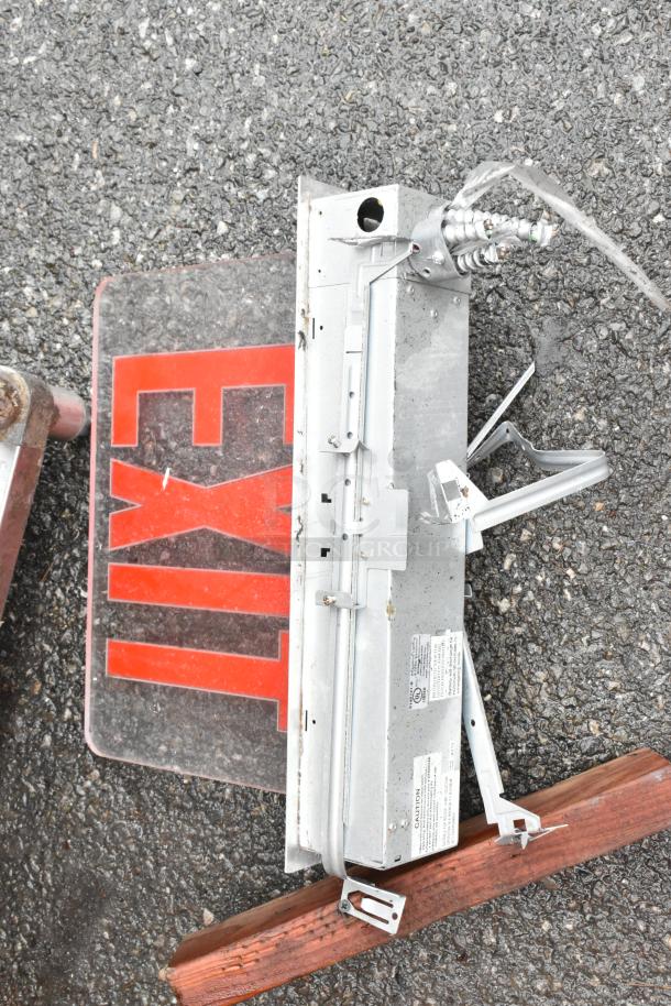 Ceiling-mounted exit sign fixture, red lettering on clear background, with wiring. Metal brackets and caution label visible.