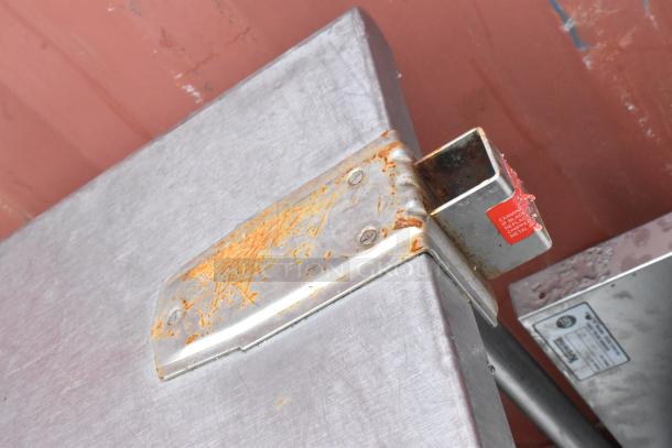 Rusty stainless steel table edge with metal bracket, a red inspection warning label, and visible wear.