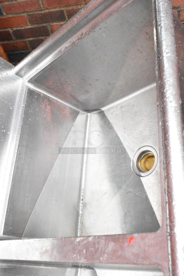 Stainless steel commercial sink with drain, showing water spots and minor wear, set against a brick wall.