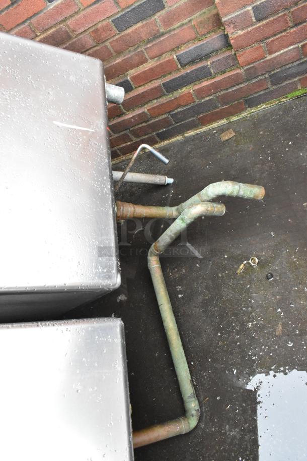 Stainless steel tank with attached copper pipes outdoors, wet surface; used condition, potential for industrial use.