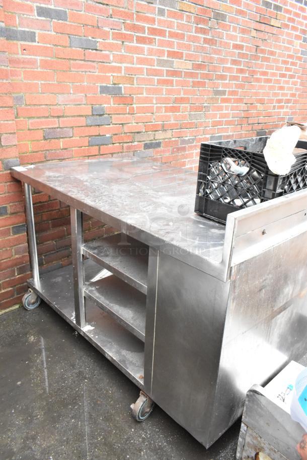 Stainless steel commercial work table with wheels, shelves, and storage compartments; moderate wear; ideal for kitchen use.