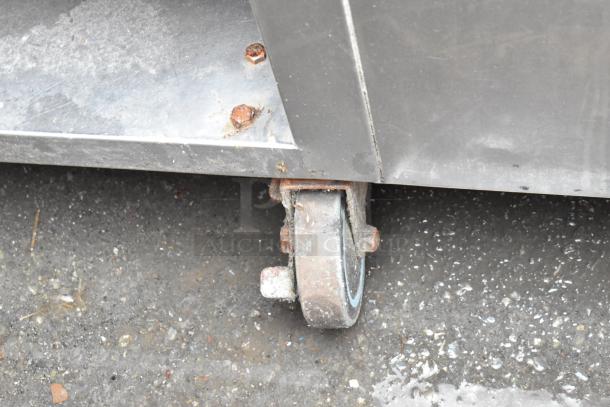Rusty metal caster wheel attached to a stainless steel frame. Visible wear and rust on wheel and bolts, indicating aged condition.