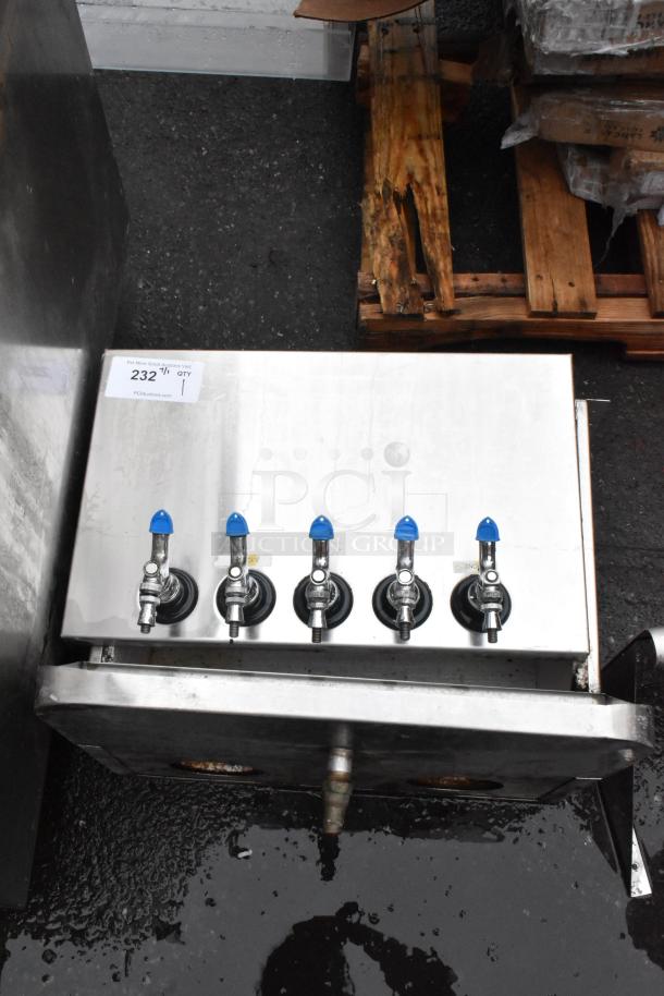 Stainless steel beverage dispenser with five blue-handled taps, marked "232," resting on a wooden pallet.