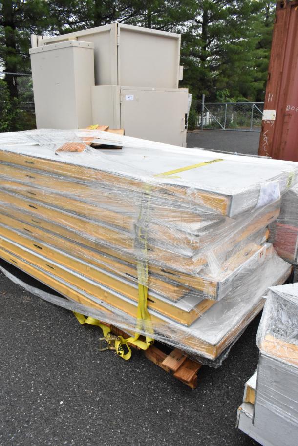 Stack of insulated metal panels wrapped in plastic with yellow straps, on a wooden pallet, outdoor setting.
