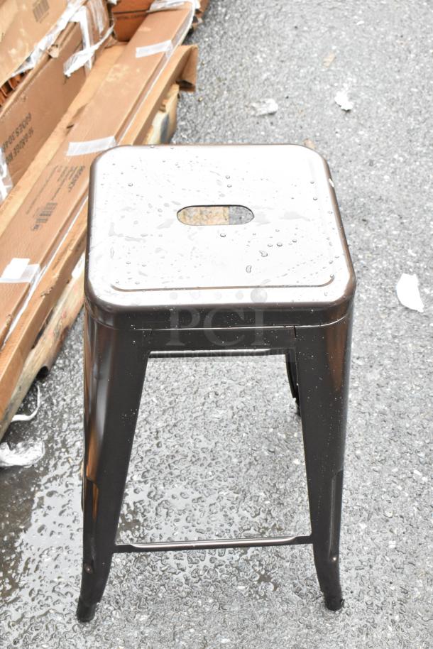 Industrial metal stool, square top with cutout handle, glossy black finish, minor wear, outdoor setting, no visible markings.