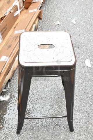 Industrial metal stool, square top with cutout handle, glossy black finish, minor wear, outdoor setting, no visible markings.