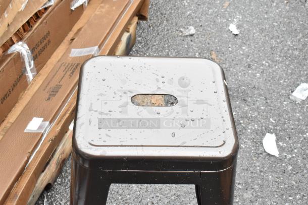 Metal stool with a rectangular seat and center handle, wet from rain, on a street with packaging boxes nearby.