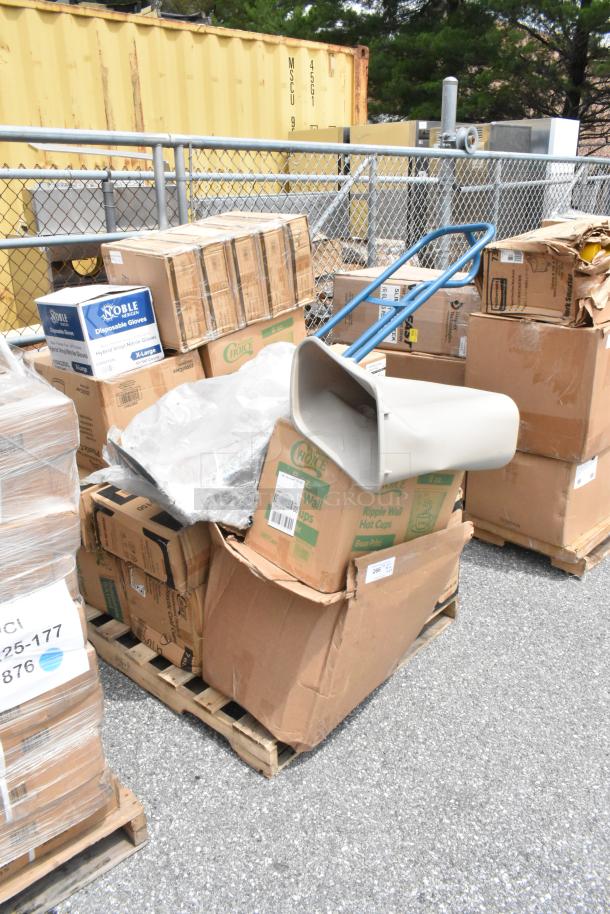 Large stack of mixed cardboard boxes and packaging on pallets, containing various items, including a plastic cart handle. Boxes show brand logos like "Noble" and "Choice," indicating disposable gloves and hot cups. Condition appears varied, with some boxes slightly damaged. Items are outdoors beside a yellow shipping container and metal fence.