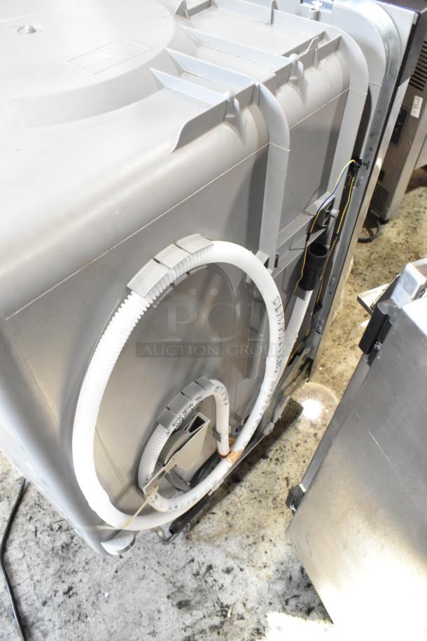 Side view of a gray dishwasher in used condition, showing hose connections and various internal components.