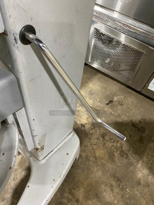 Metallic lever on industrial machine, with visible wear and scratches, affixed to a gray metal base on concrete floor.