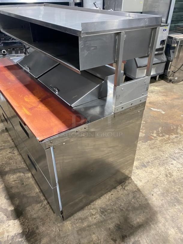Stainless steel commercial kitchen prep station with wood cutting board, upper shelf, and multiple compartments. Good condition.