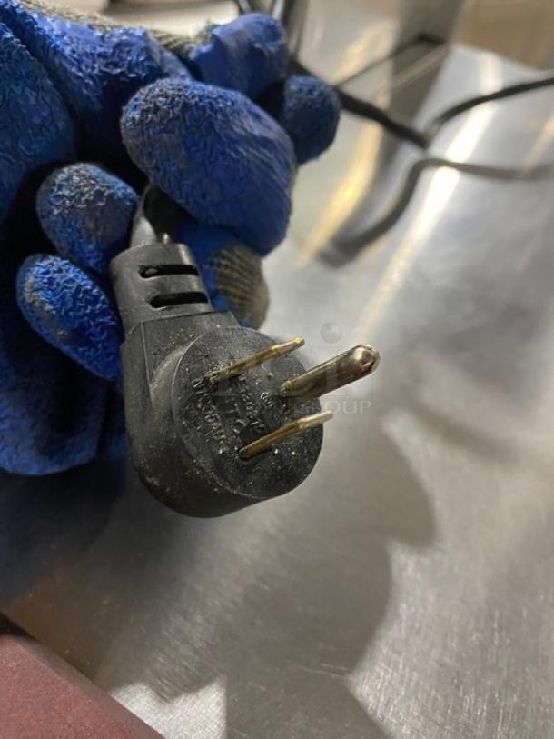 Black electrical plug with three prongs, held by blue-gloved hand. Surface shows signs of wear, marking indicates 250V.
