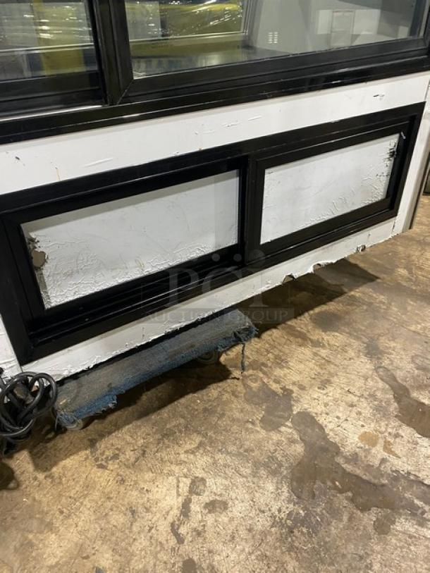 Commercial display cooler with black frame, showing wear and peeling paint on the lower panels. Power cord visible.