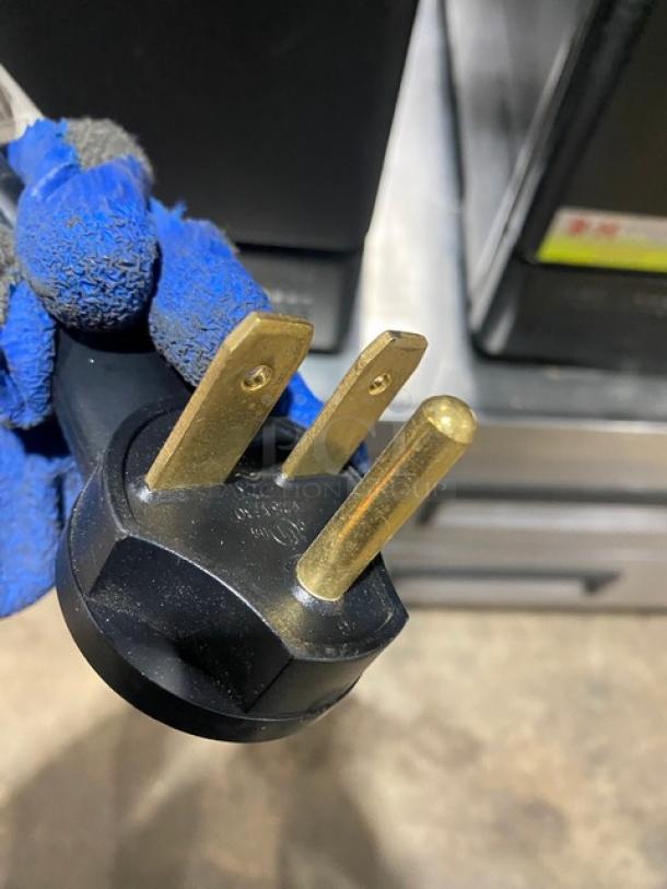 Close-up of a three-prong electrical plug with visible wear, held by a gloved hand, showing standard gold blades.