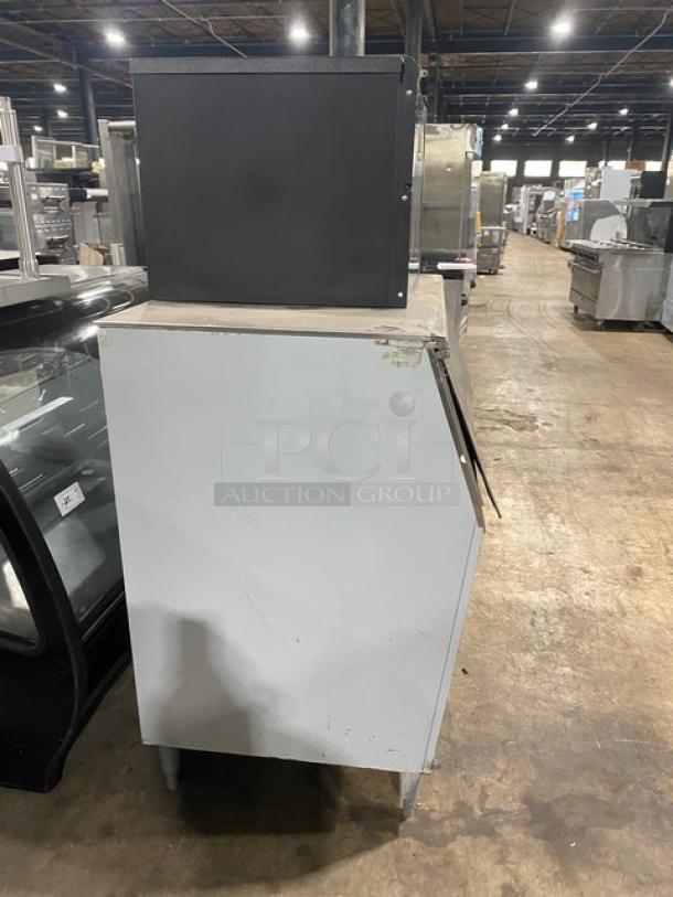 Commercial ice maker machine with black top unit, showing wear on sides, in a warehouse setting. Ideal for restaurant use.