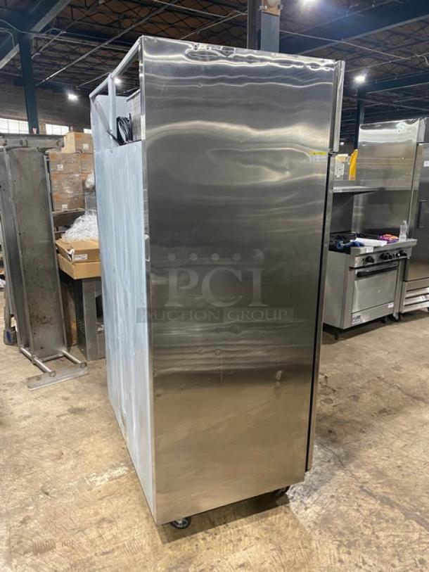 Commercial stainless steel refrigerator on wheels, side view. Appears in good condition, with a single door design. Industrial setting.