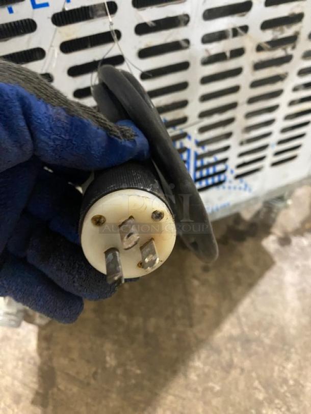 Heavy-duty electrical plug with three prongs and black cable. Gloved hand holding it. Signs of wear. Background shows vented metal surface.
