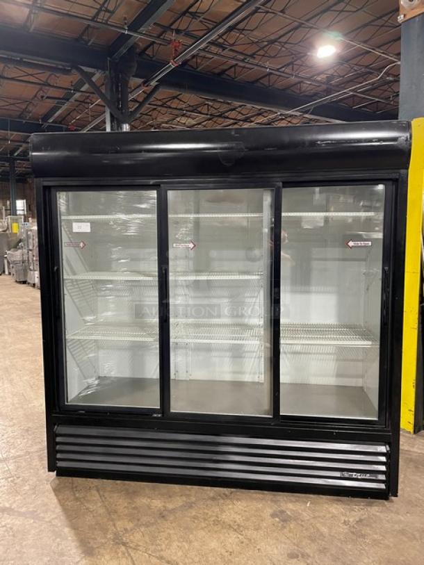 Black three-door commercial glass display refrigerator with multiple wire shelves, in a warehouse setting. Good condition.