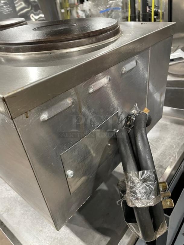 Stainless steel industrial hot plate with dual burners, power cables attached. Signs of wear on surfaces. Ideal for heavy-duty use.