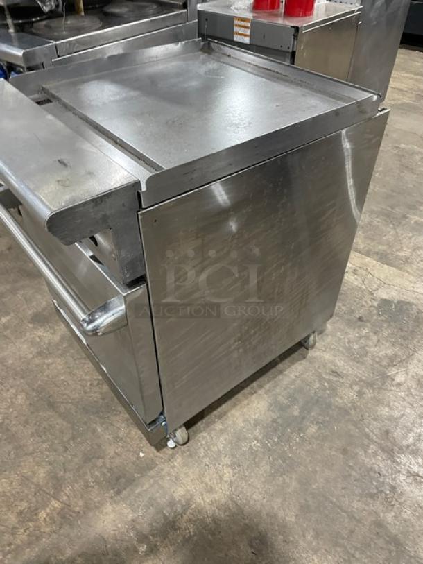 Stainless steel commercial flat griddle on wheels, with dual storage compartments. Some surface wear visible.