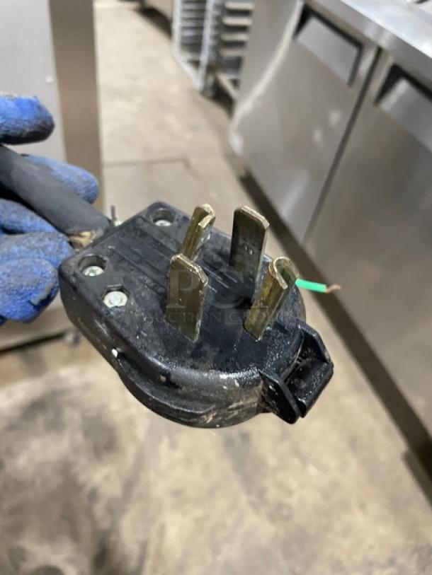 Three-prong industrial power plug, black casing, metal prongs, slight wear, exposed green wire, suitable for heavy-duty equipment.
