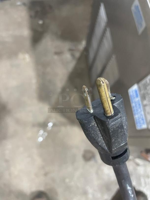 Worn black electrical plug with visible metal prongs, showing signs of use. Background labels are blurred and unreadable.