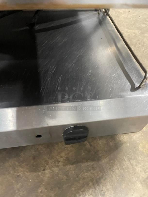 Industrial griddle with stainless steel surface, visible wear marks, metal rail, and small handle, used condition.
