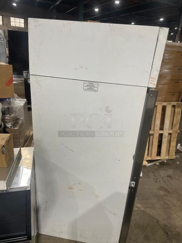 Commercial upright refrigerator, white exterior, visible wear, metal door handle. Label indicates model details, industrial use.