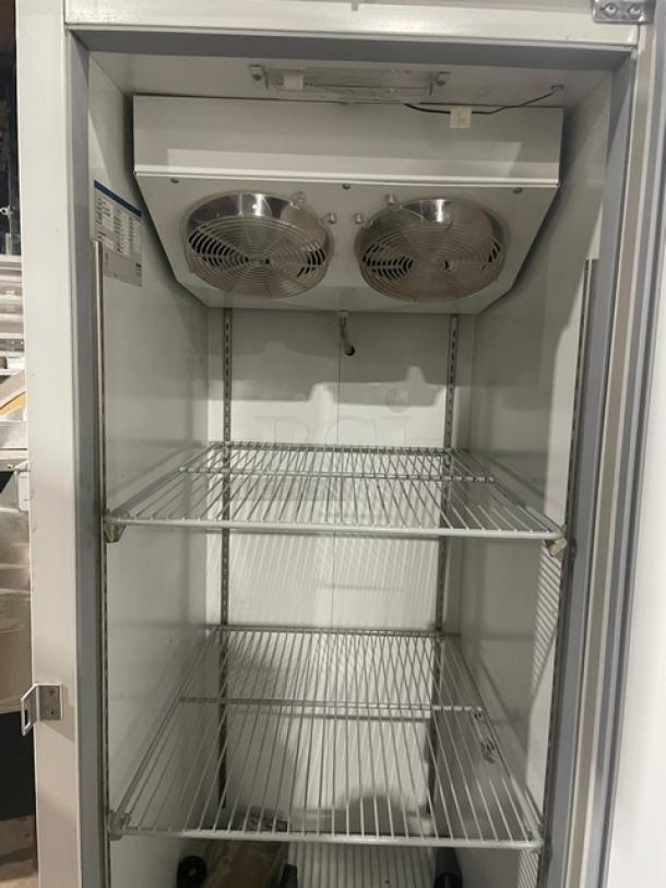 Commercial single-door refrigerator with two wire shelves and twin internal cooling fans. Clean interior, stainless steel finish, label on side panel.
