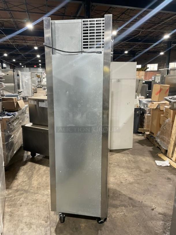 Tall stainless steel commercial refrigerator on wheels, vented top, minor wear, suitable for industrial kitchen use.
