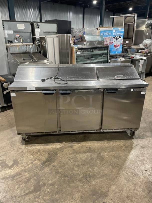 Stainless steel commercial sandwich prep table with two refrigerated compartments, three hinged lids, and wheeled base.