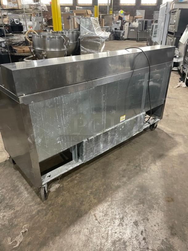 Stainless steel commercial refrigerated prep table on wheels, visible wear, includes ventilation unit, power cord attached.