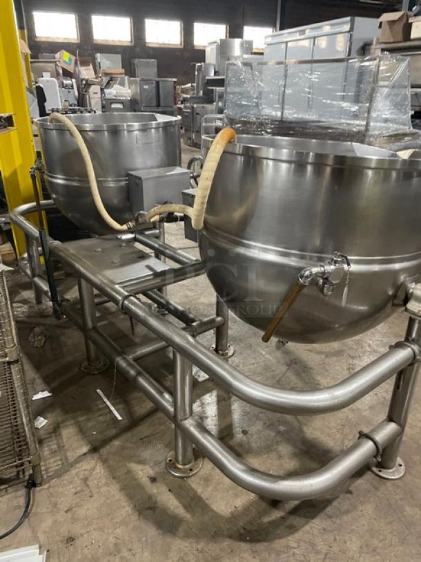 Industrial stainless steel dual steam kettles with support frame, used condition, featuring control valves and attached hoses.