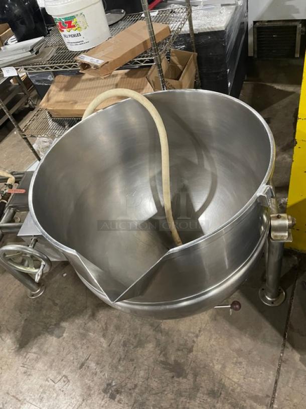 Industrial stainless steel tilt kettle with spout and attached hose, on sturdy legs. Good condition, ideal for commercial use.