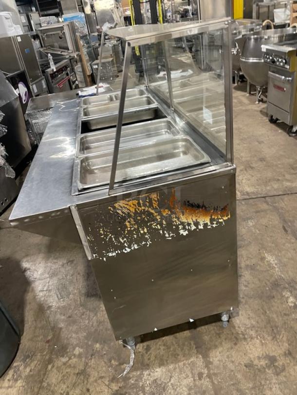 Commercial stainless steel serving counter with four food trays and a glass sneeze guard. Visible paint wear on the side.