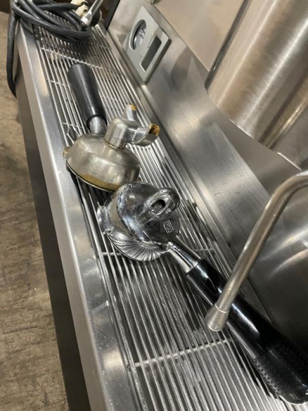 Commercial espresso machine portafilters, showing wear. One with dual spouts, the other single. Set on a metal drip tray.
