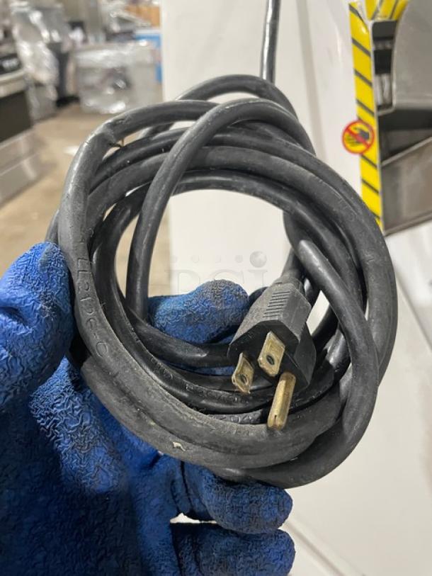 Coiled black power cord with a two-prong plug, held by a gloved hand. Cord appears in good condition.