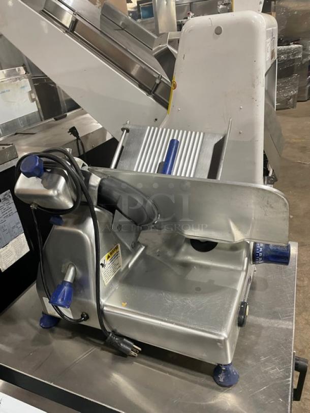 Commercial deli meat slicer, stainless steel, with blue handles and power cord, in good condition, on a metal surface.