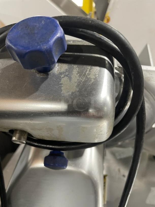 Commercial meat slicer with silver body, blue adjustment knobs, and visible wear. Includes black power cord.