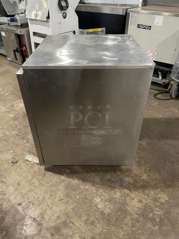 Stainless steel commercial fridge, box-style, used condition with visible scuffs. No visible brands or labels.