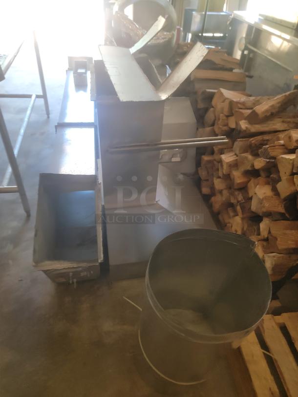 Metal industrial meat grinder with hopper and loading tray, in used condition, set by stacked wood. No visible markings.