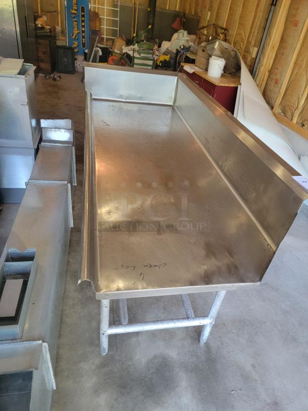 Stainless steel worktable with raised edges, in used condition. Handwritten markings on surface, sturdy metal legs.