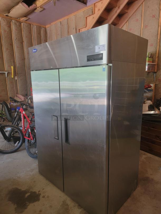 Stainless steel two-door commercial refrigerator, brand "Arctic," in used condition with visible label, located in garage setting.