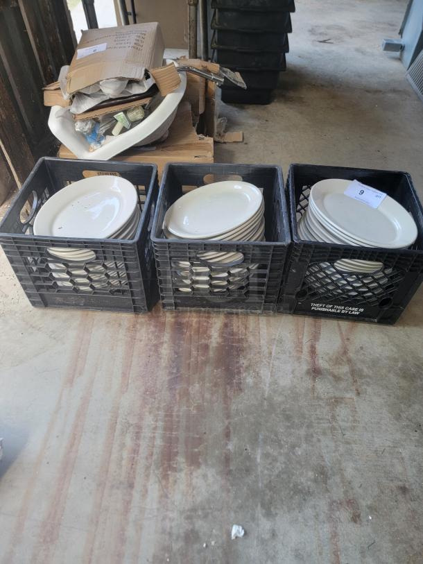Three crates of white ceramic plates in used condition, tagged for auction, with an additional cardboard box of miscellaneous items in the background.