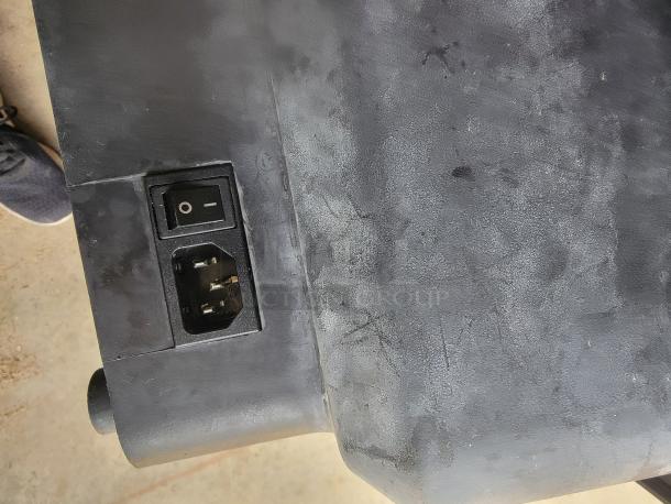 Black electronic device with power switch and three-prong plug socket on textured surface, showing wear and scratches.